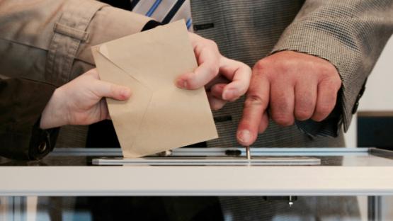 Image of three hands voting, casting a ballot paper into a box together.