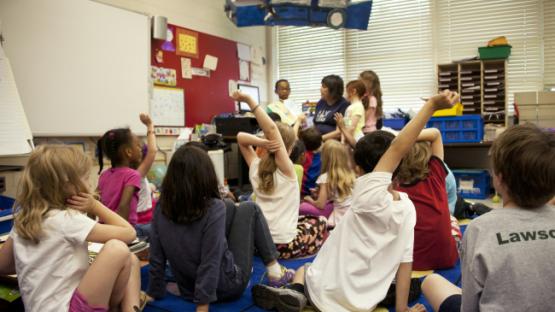 typical classroom scene, where an audience of school children were seated on the floor before a teacher at the front of the room, who was reading an illustrated storybook, during one of the scheduled classroom sessions.