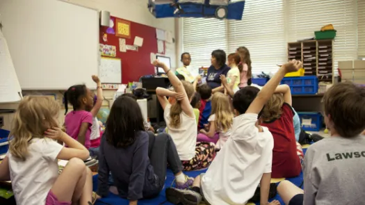 typical classroom scene, where an audience of school children were seated on the floor before a teacher at the front of the room, who was reading an illustrated storybook, during one of the scheduled classroom sessions.