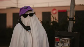 A person in a white sheet, representing a ghost, wearing a purple hat that says spooky. On a payphone outside in the street